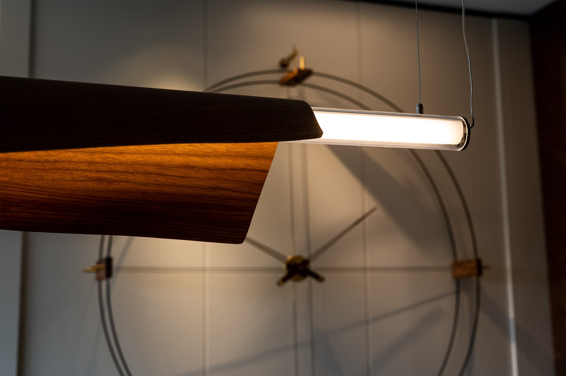 Folded walnut linear pendant with a glowing tube lit before an open wire wall clock in Timeless Union