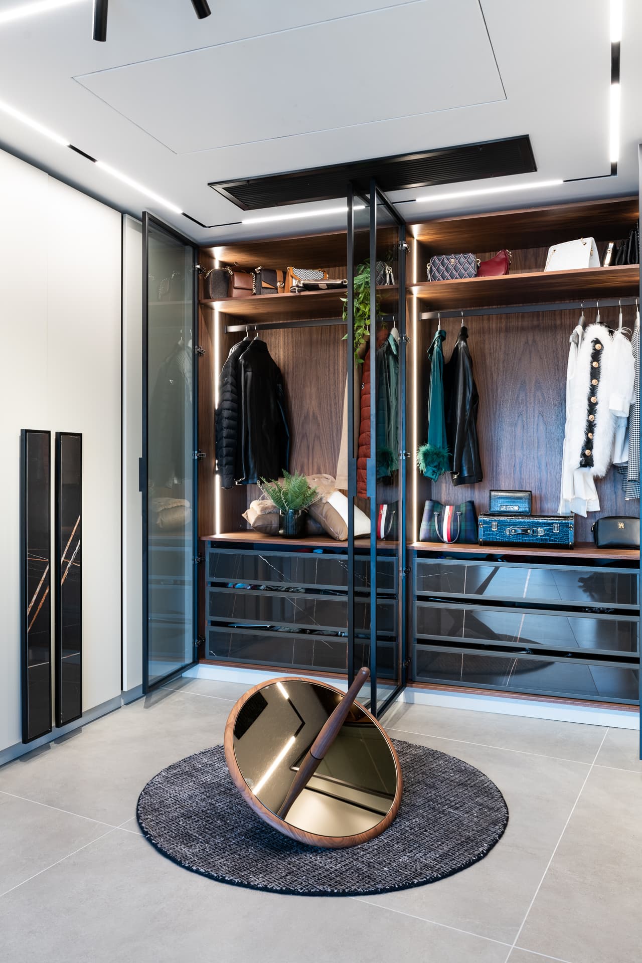 Walk-in dressing room with glass doors, lit walnut shelving and a tilted round floor mirror on a round rug in Timeless Union