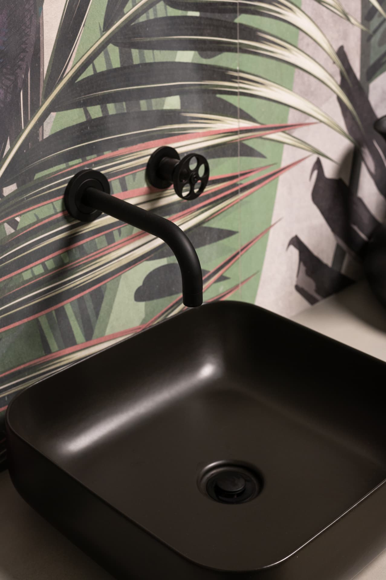 Matte black square basin with a wall spout and wheel tap on a tropical palm wallcovering in the guest bathroom of Timeless Union