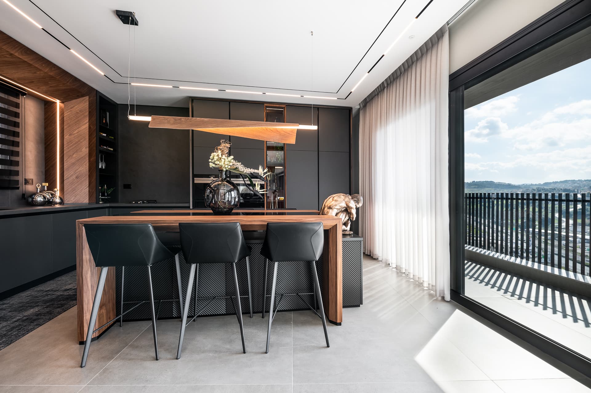 Walnut kitchen island with black leather bar stools under a folded wood linear pendant, beside a glass wall in Timeless Union