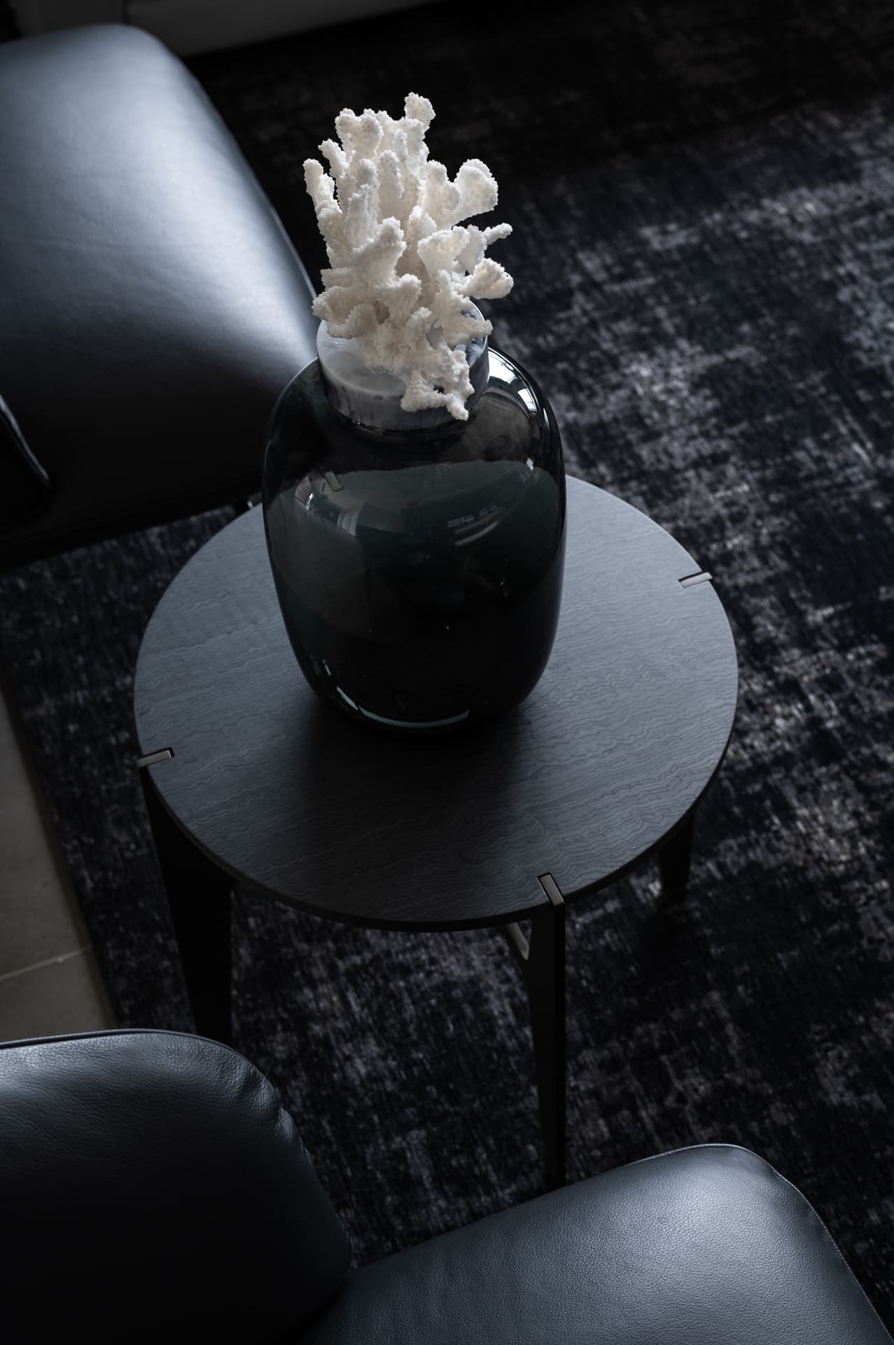 White coral on a smoked glass vase atop a round black side table between leather armchairs in the living room of Timeless Union