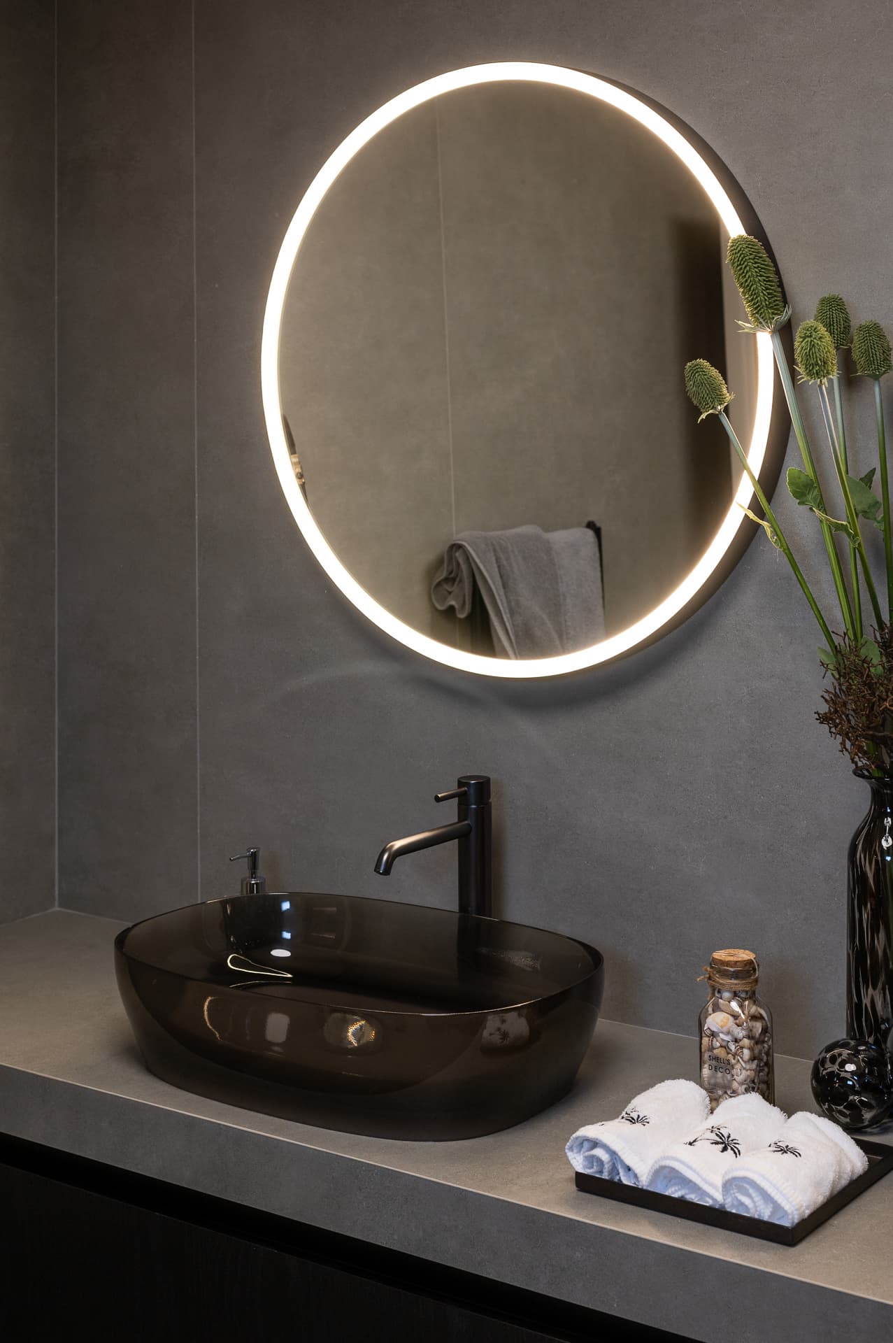 Smoked glass vessel basin under a backlit round mirror on a grey stone counter in the master bathroom of Timeless Union