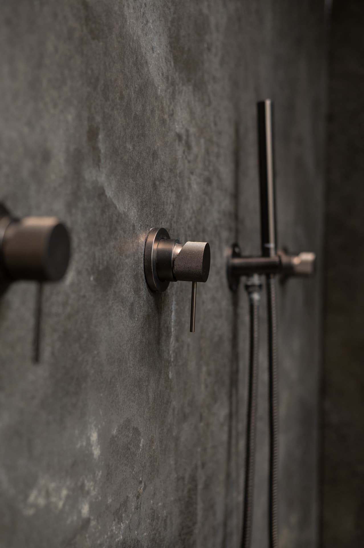 Knurled dark bronze shower valves and hand shower on a textured grey stone wall in the master bathroom of Timeless Union