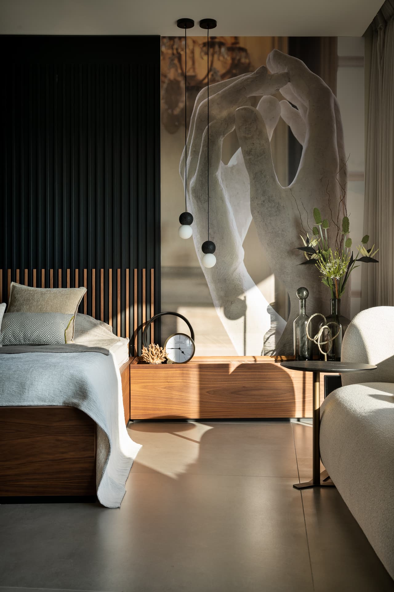 Low walnut bedside unit with a round clock beneath twin orb pendants and a sculpted hands artwork in the master bedroom of Timeless Union