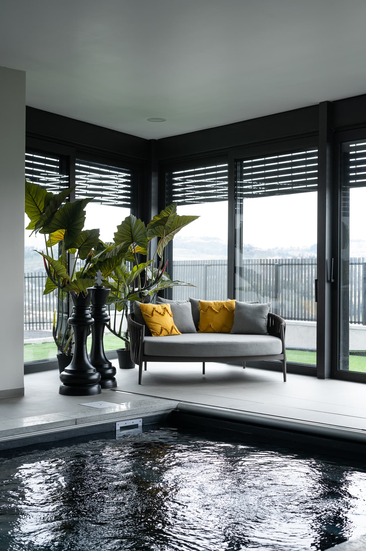 Indoor pool edge with a grey rope daybed, mustard cushions and oversized black chess pieces by glass walls in Timeless Union