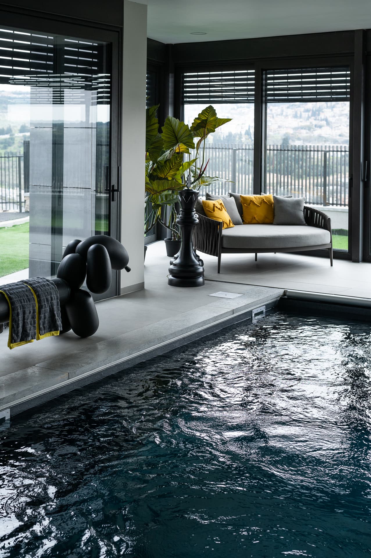 Indoor pool with a black balloon-dog sculpture draped with a towel, a giant chess piece and corner daybed by glass walls in Timeless Union