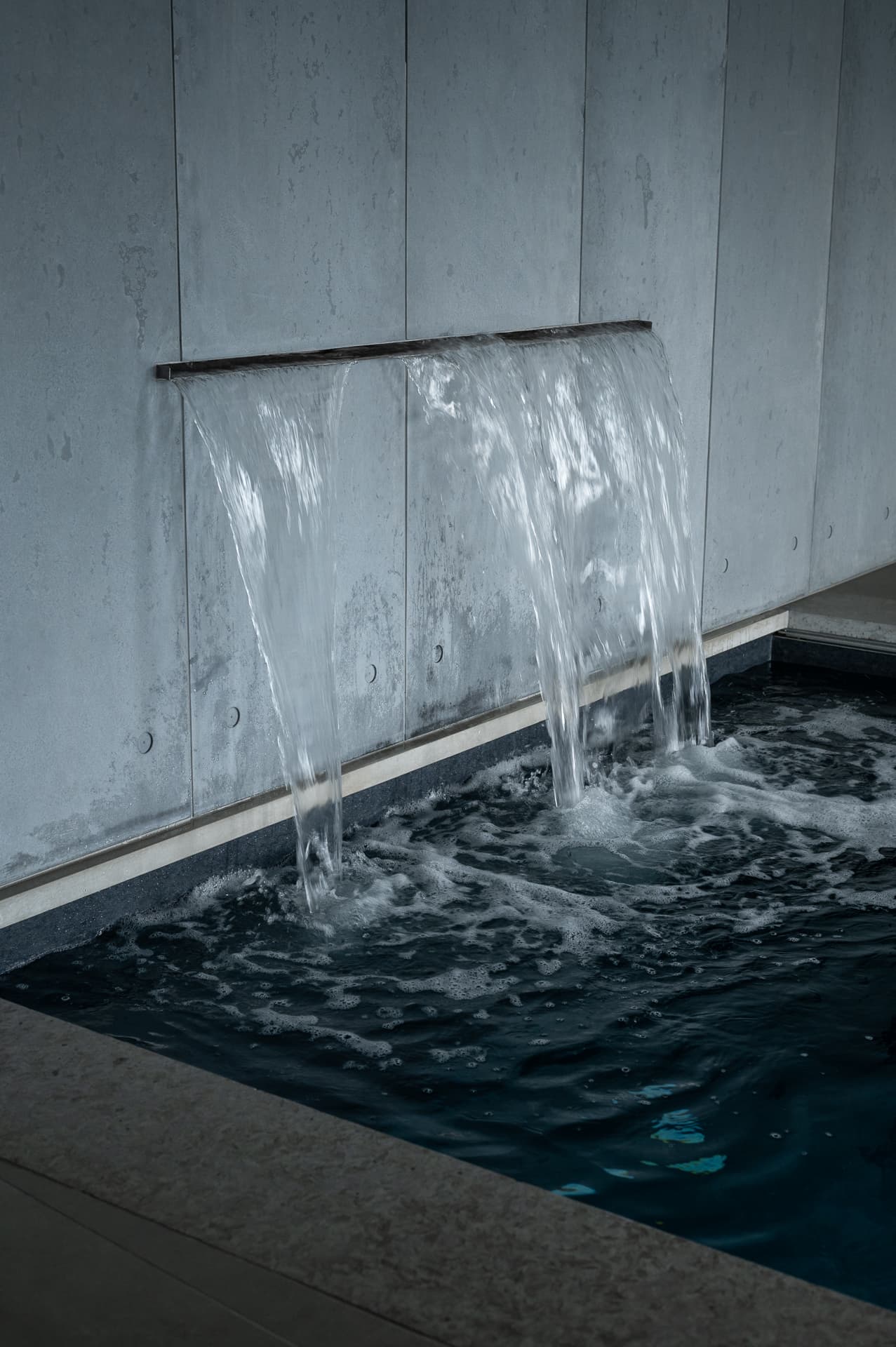 Sheet water spout pouring from a board-formed concrete wall into the dark pool in Timeless Union