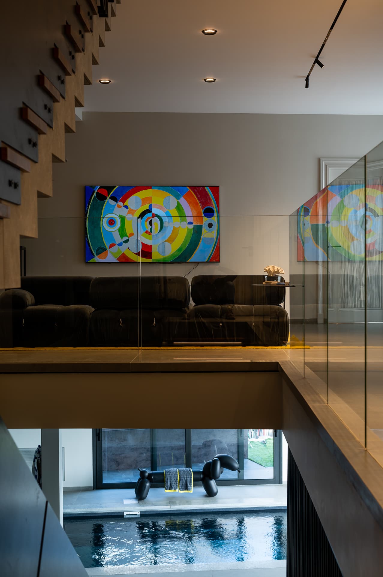 Glass-railed gallery lounge with a dark modular sofa and colourful circular artwork above the pool in Timeless Union