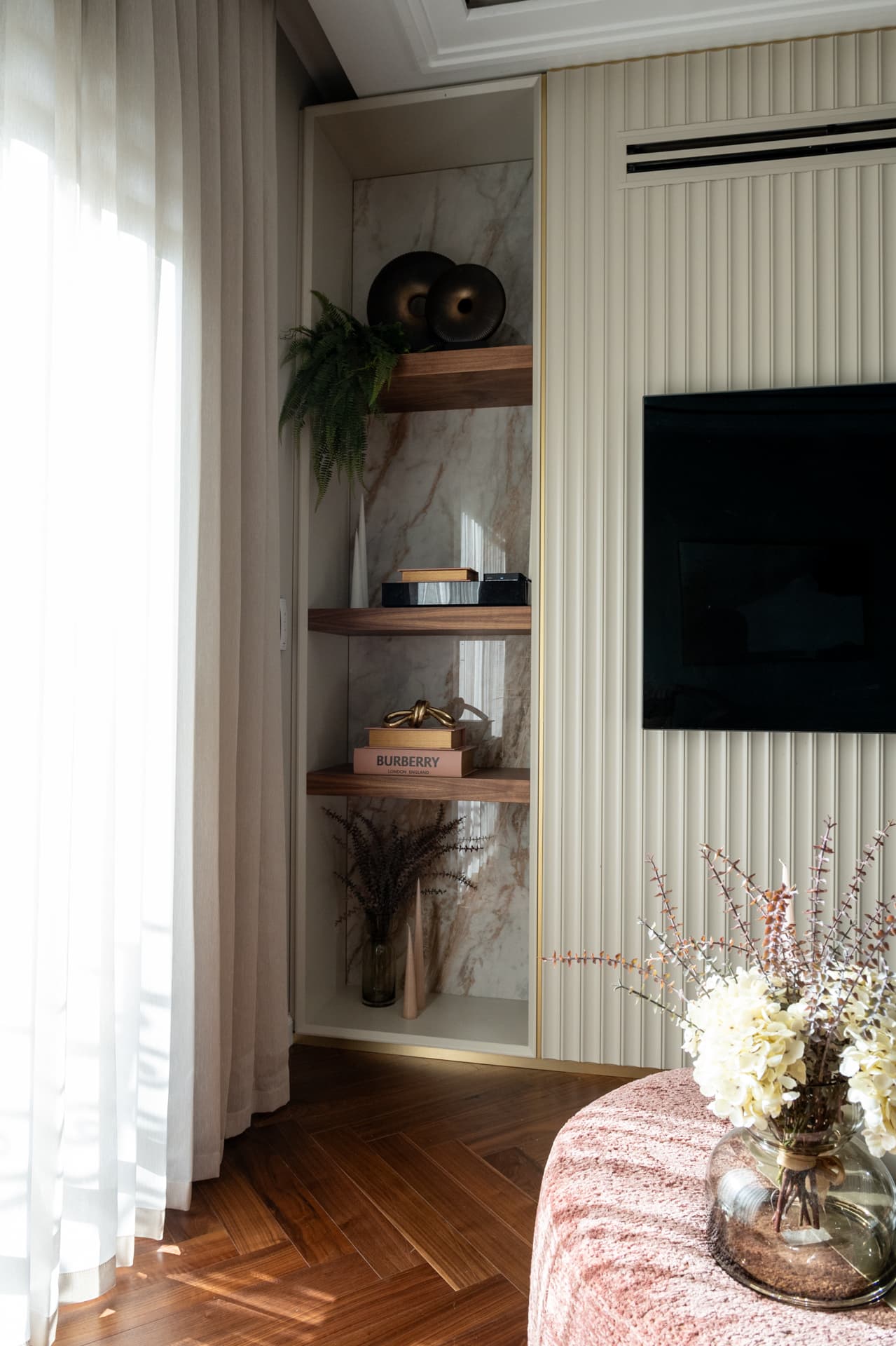 Walnut floating shelves in a veined stone niche beside a fluted cream wall panel and a blush round ottoman in the second bedroom of Twilight