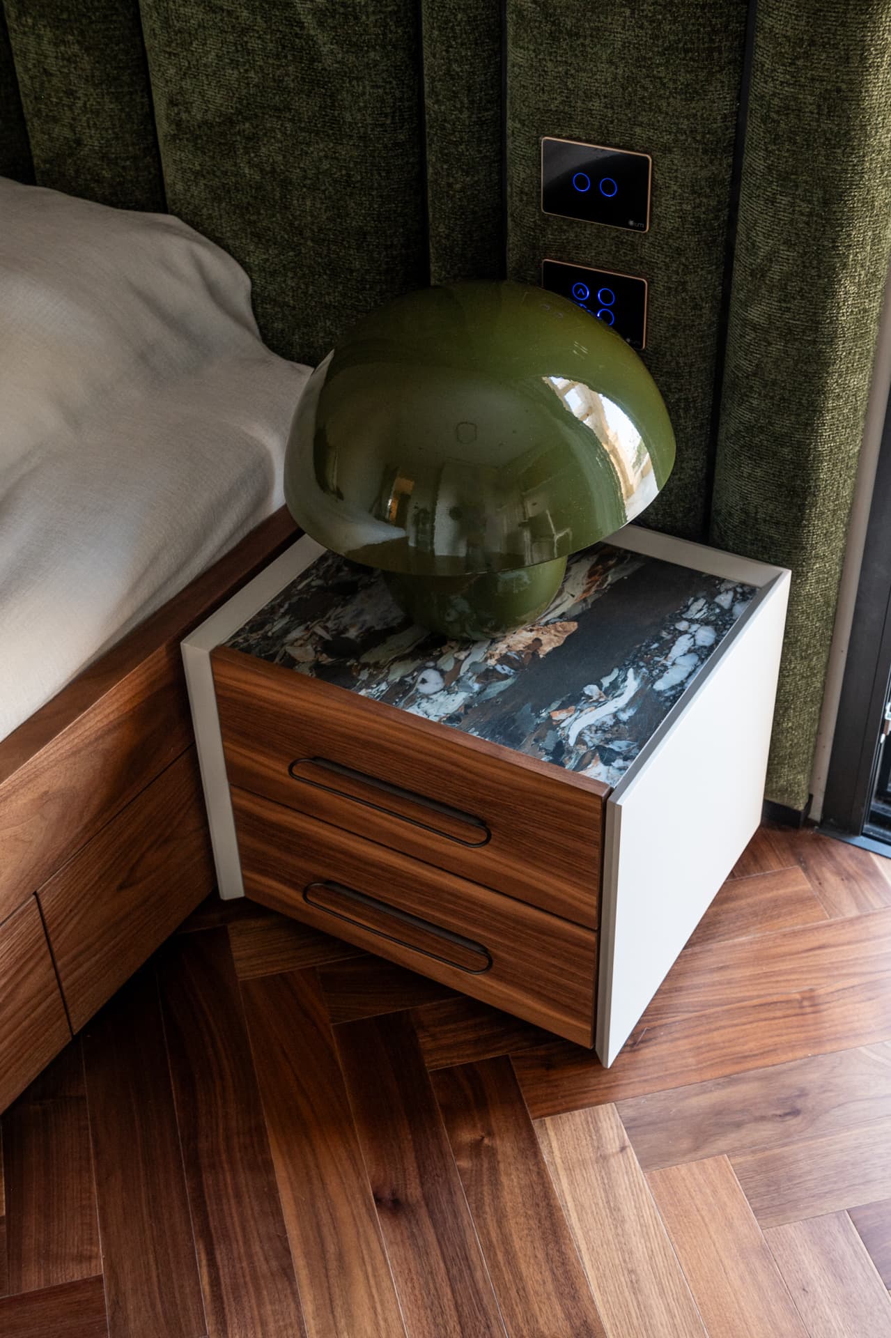 Green glass mushroom lamp on a walnut nightstand with a dark stone top by a moss green padded headboard in the third bedroom of Twilight