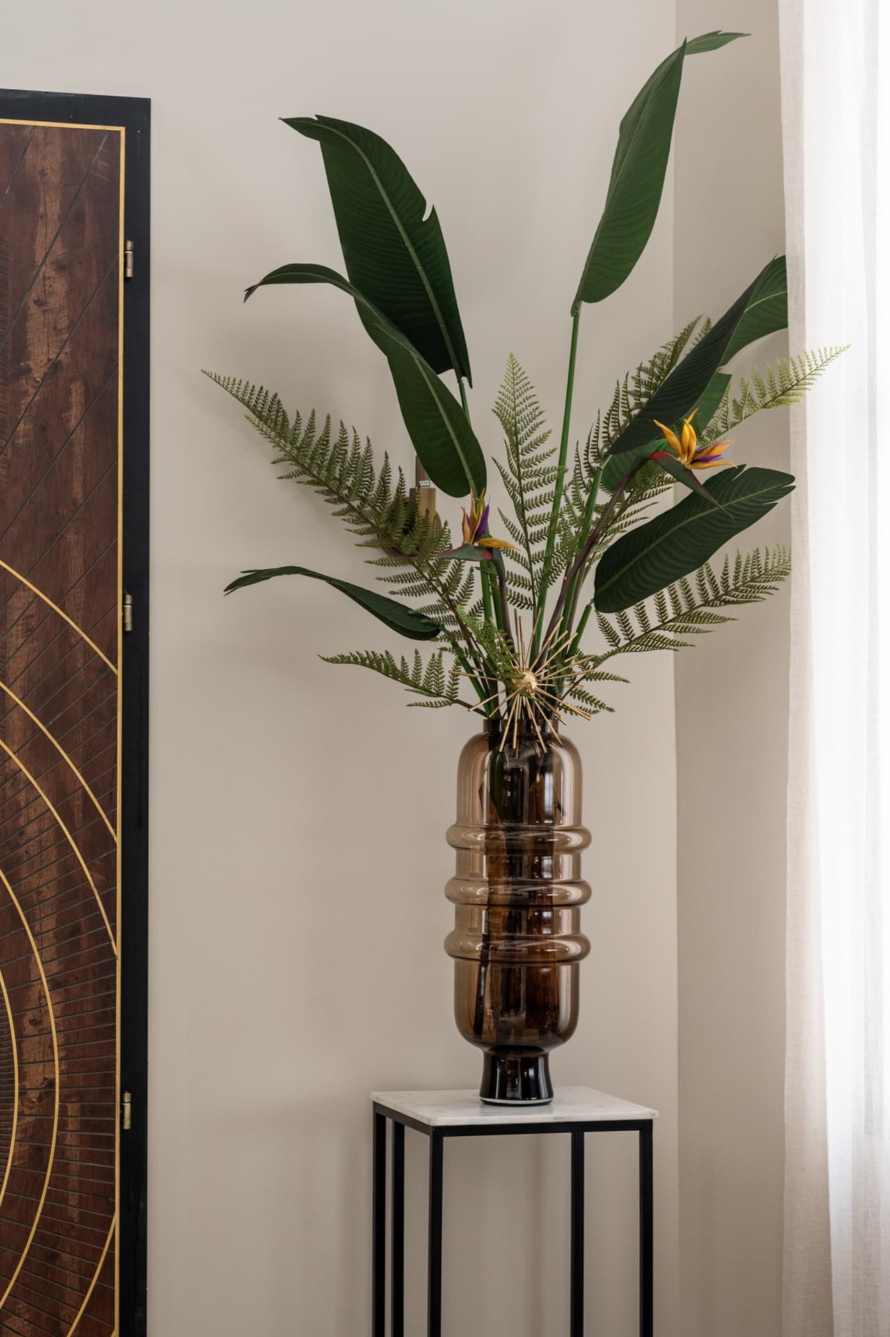 Tall ribbed amber glass vase with bird-of-paradise leaves and ferns on a stone-topped stand beside a brass-inlaid screen in Twilight