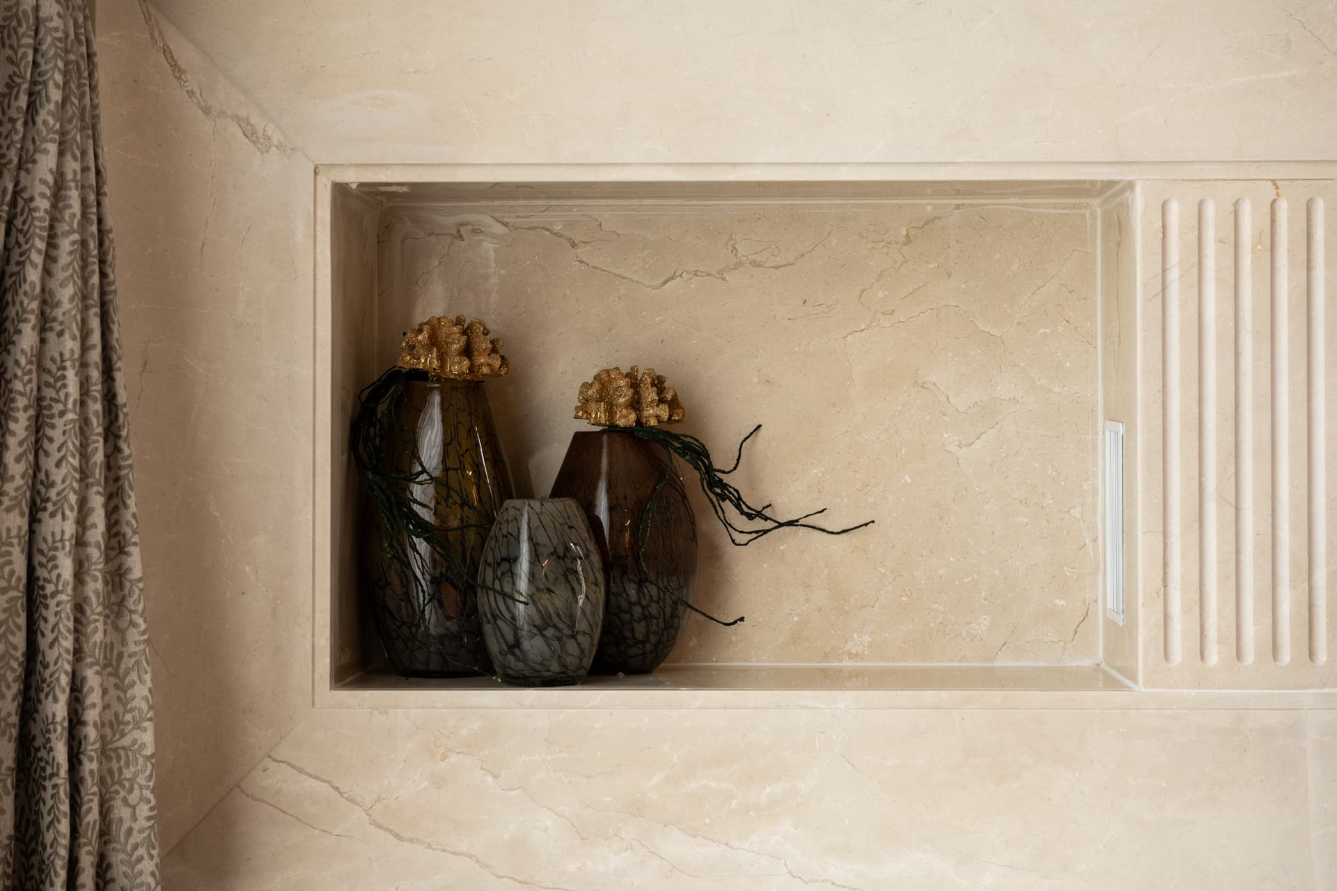 Recessed beige stone niche with fluted edge holding smoked glass vases topped with gilded coral forms in the entrance of Twilight