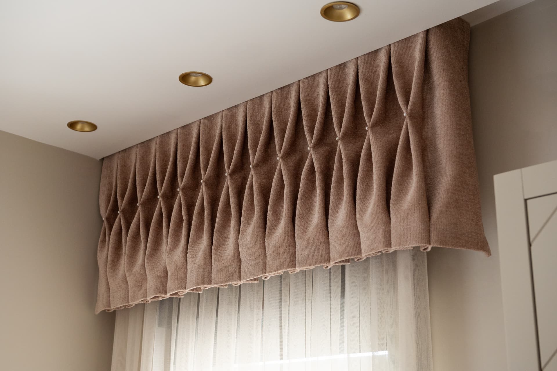 Hourglass-pleated taupe pelmet with pearl studs over sheer curtains beneath brass recessed downlights in the entrance of Twilight