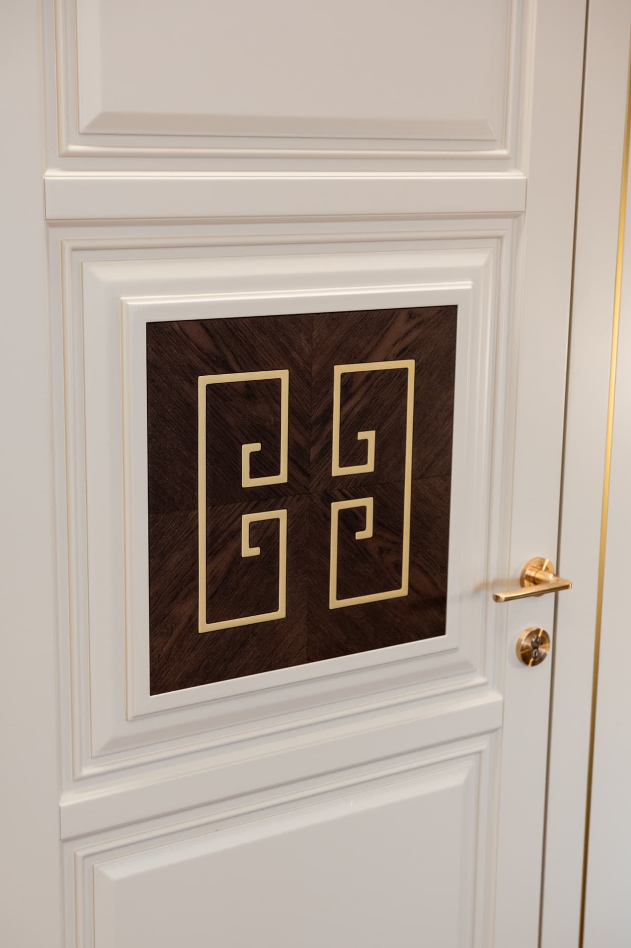 Cream panelled door with a dark wood inlay framing an interlocking brass fret motif and a brass lever handle in the entrance of Twilight