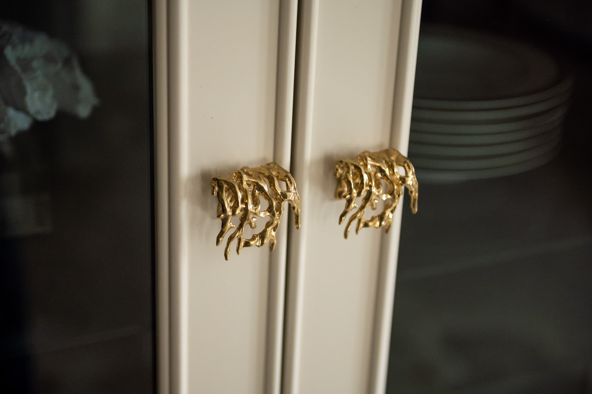 Pair of sculpted brass coral-like knobs on cream glass-fronted cabinet doors holding stacked plates in the kitchen of Twilight