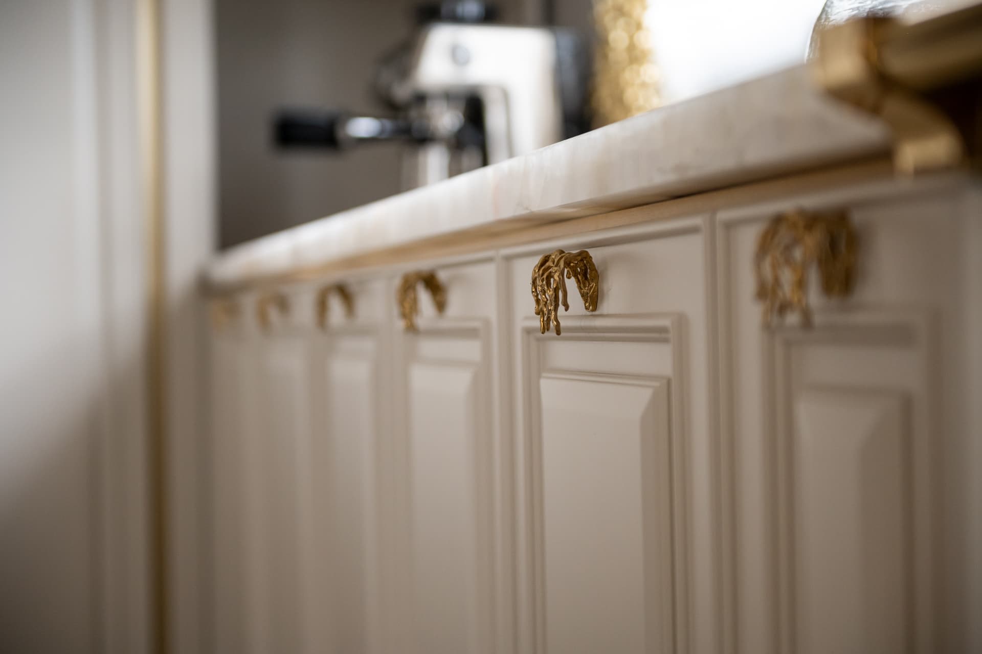 Row of cream raised-panel cabinets with sculpted brass pulls under a veined stone countertop in the kitchen of Twilight