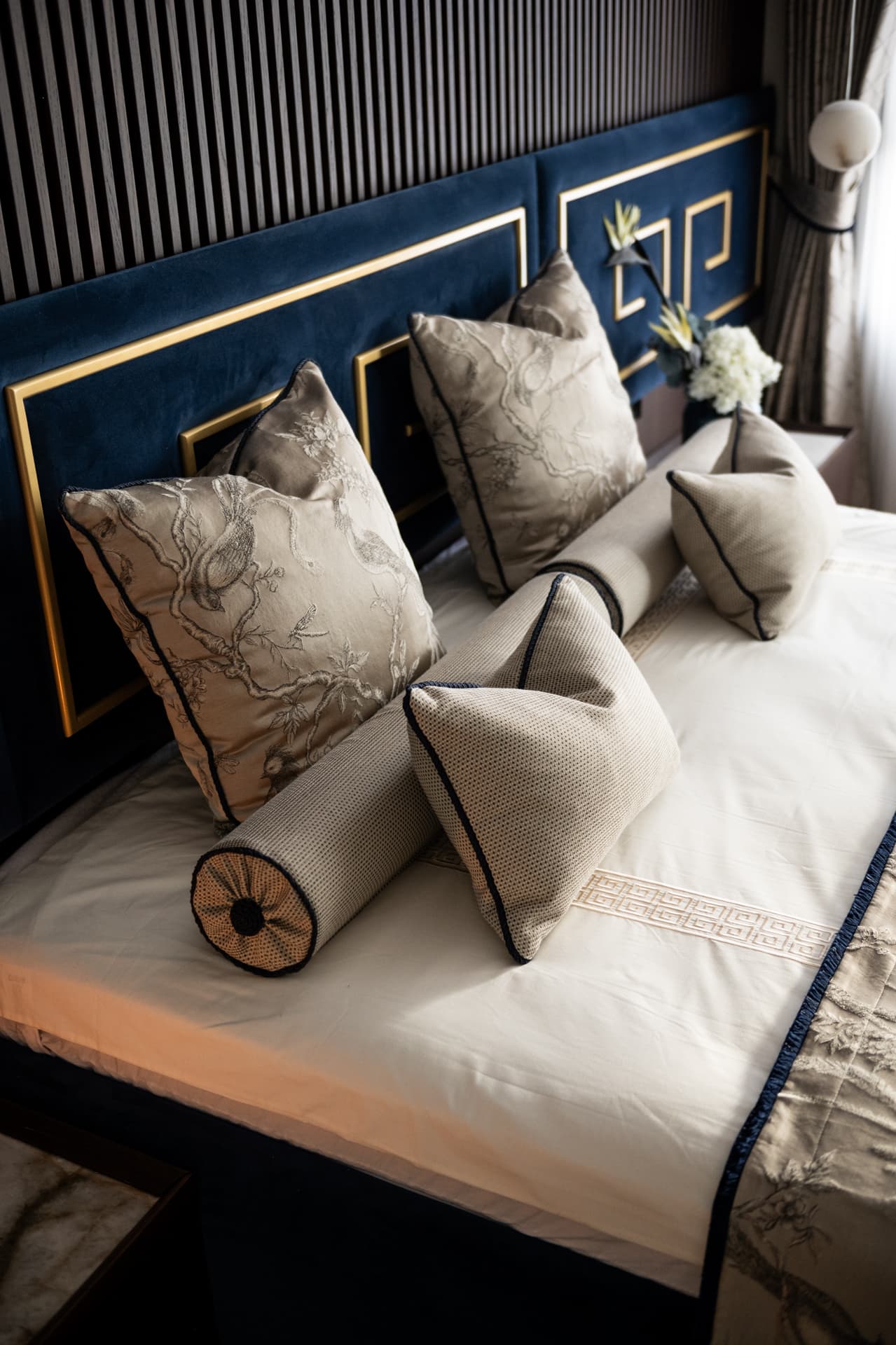 High view of piped bolsters and Greek key embroidered linen below a slatted wall and navy headboard in the master bedroom of Twilight